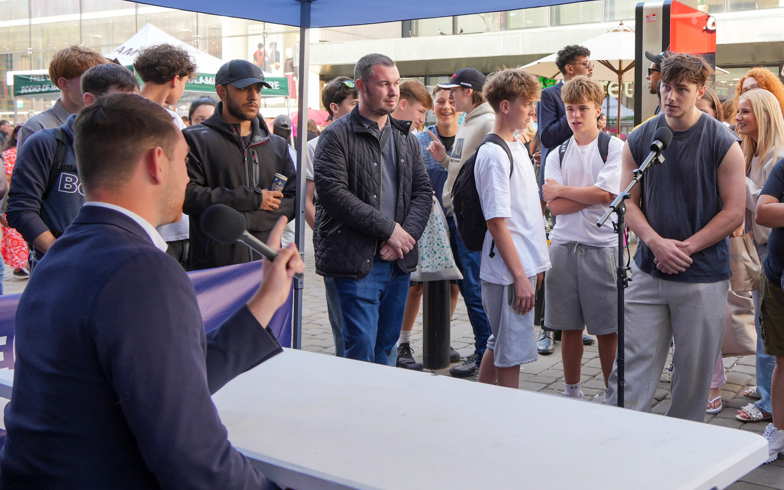 Street interview under a blue canopy — interviewer with mic facing two young men and a watching crowd