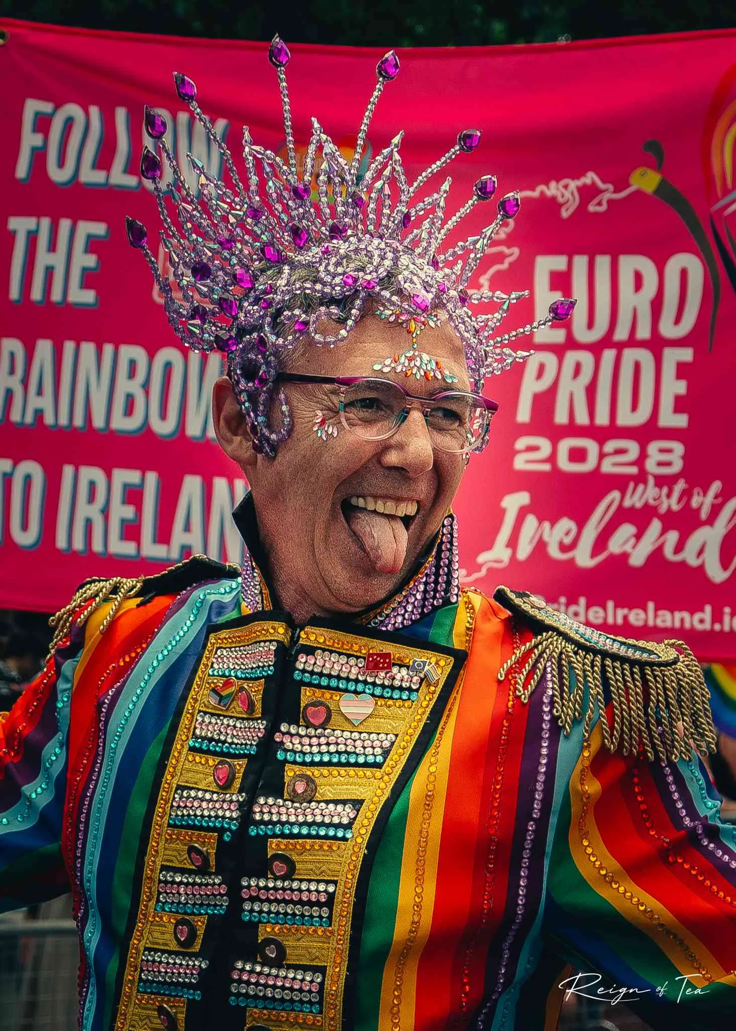 Joyful Pride portrait — rainbow military jacket and crystal headdress against a pink Euro Pride banner